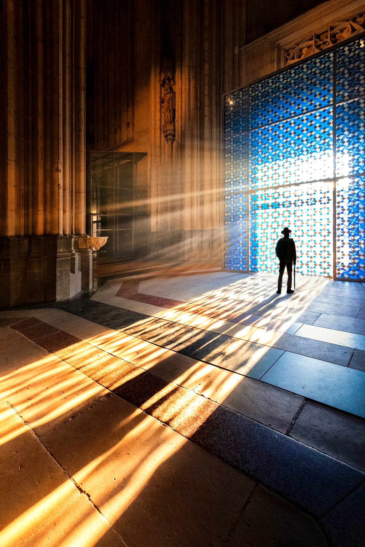 A lone figure stands before a tall blue patterned gate as sunlight casts long geometric shadows across a stone hall.