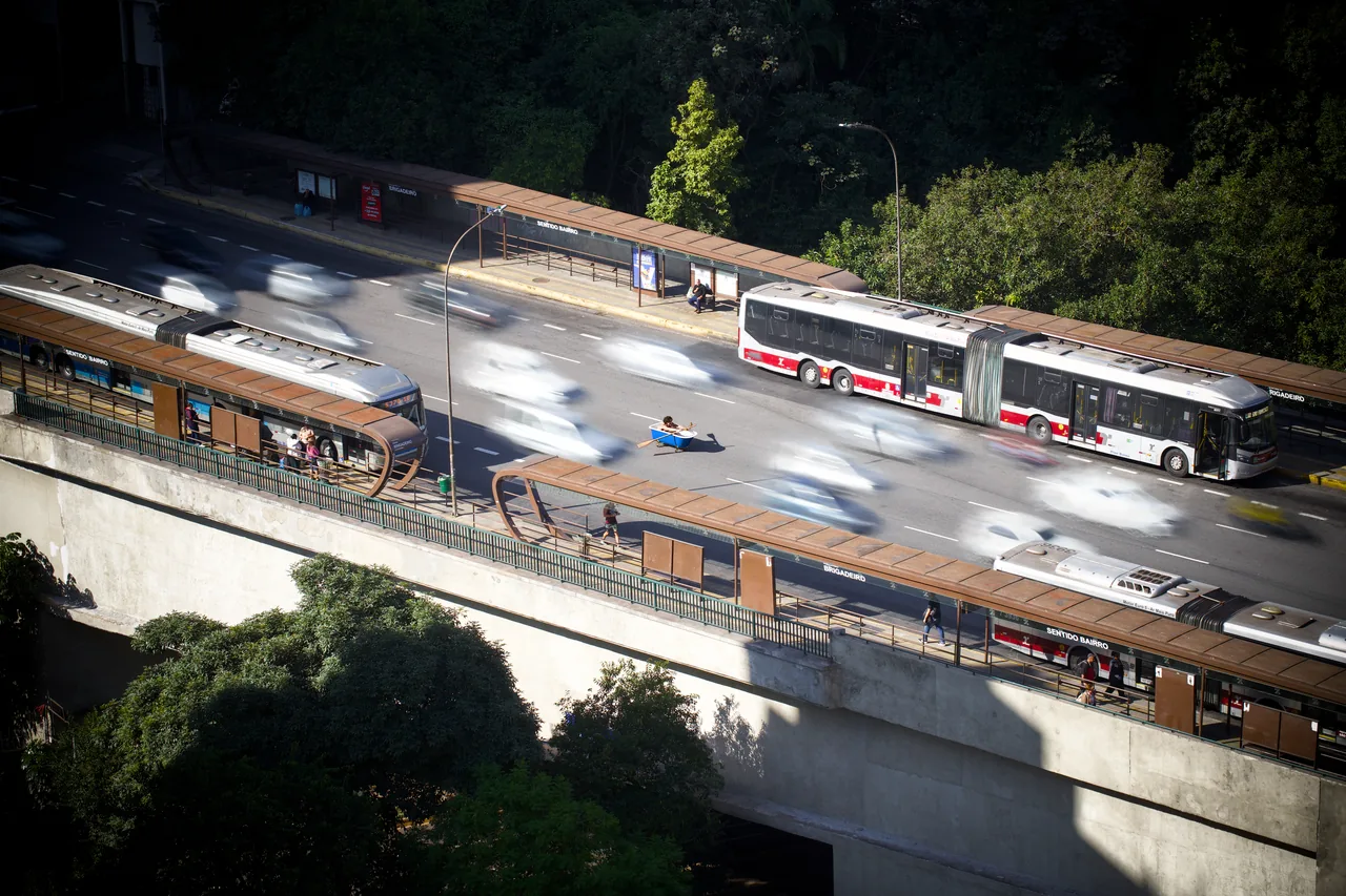 An elevated view shows blurred buses and traffic around a sharply defined person seated in a small blue tub or boat on the roadway.