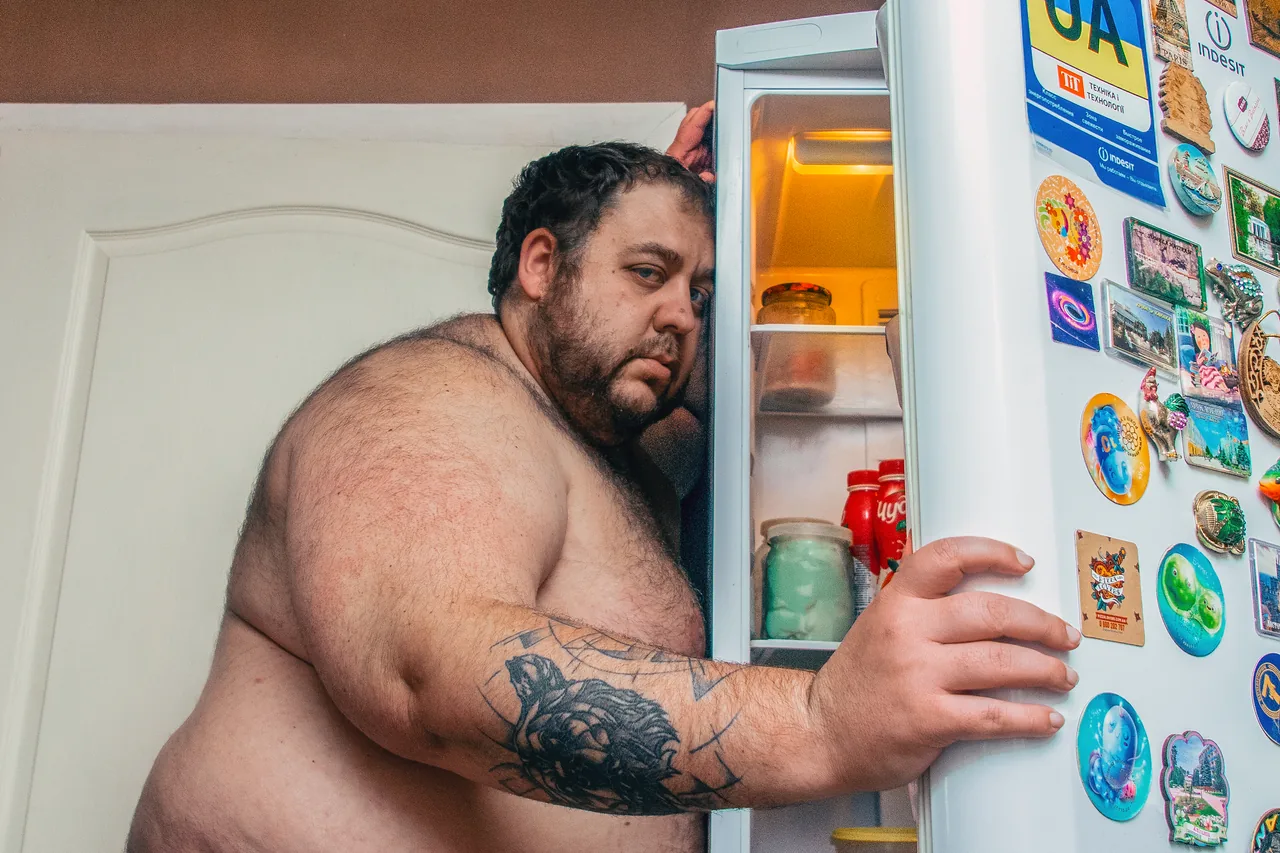 A shirtless person opens a refrigerator covered in colorful magnets in a kitchen.