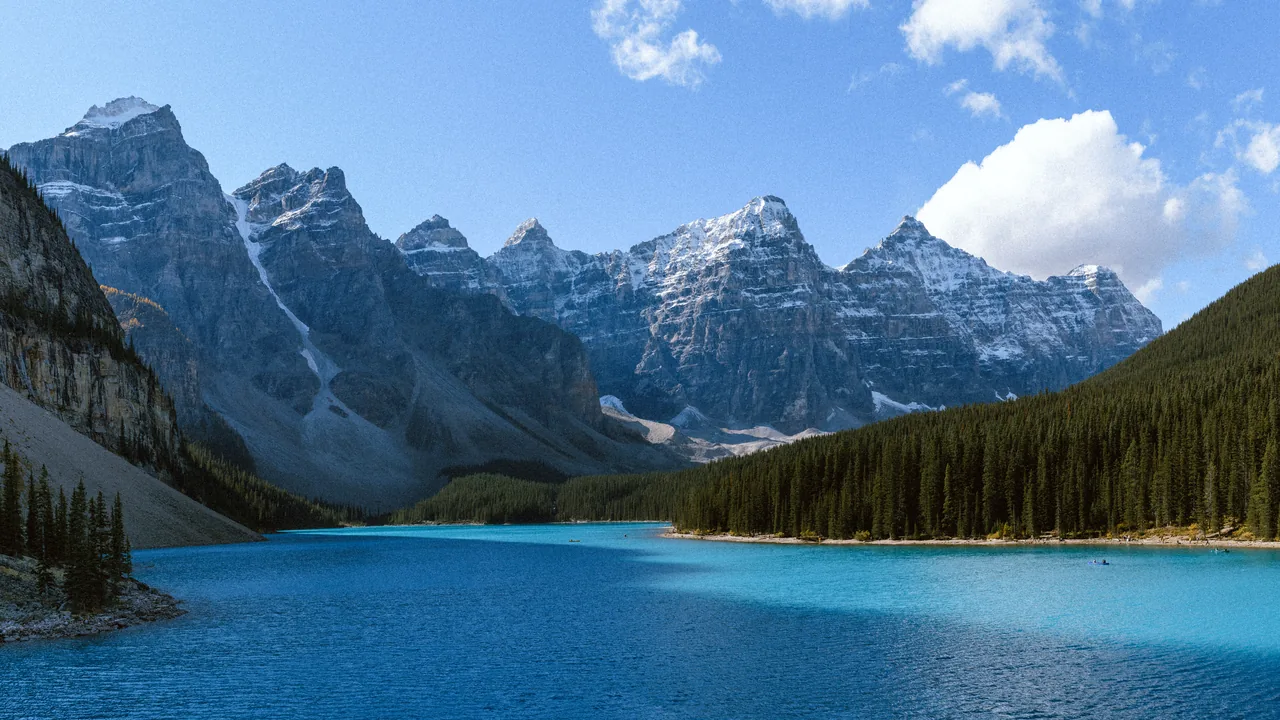 A turquoise mountain lake is bordered by evergreen forest and a jagged, snow-dusted mountain range.