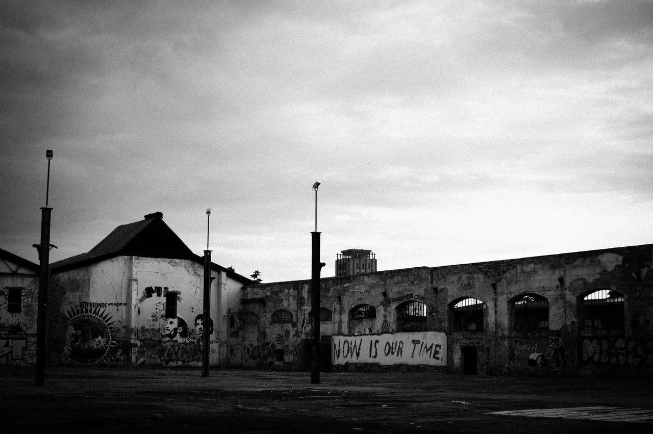 Black-and-white industrial buildings sit beneath a cloudy sky, with the words NOW IS OUR TIME painted on a wall.