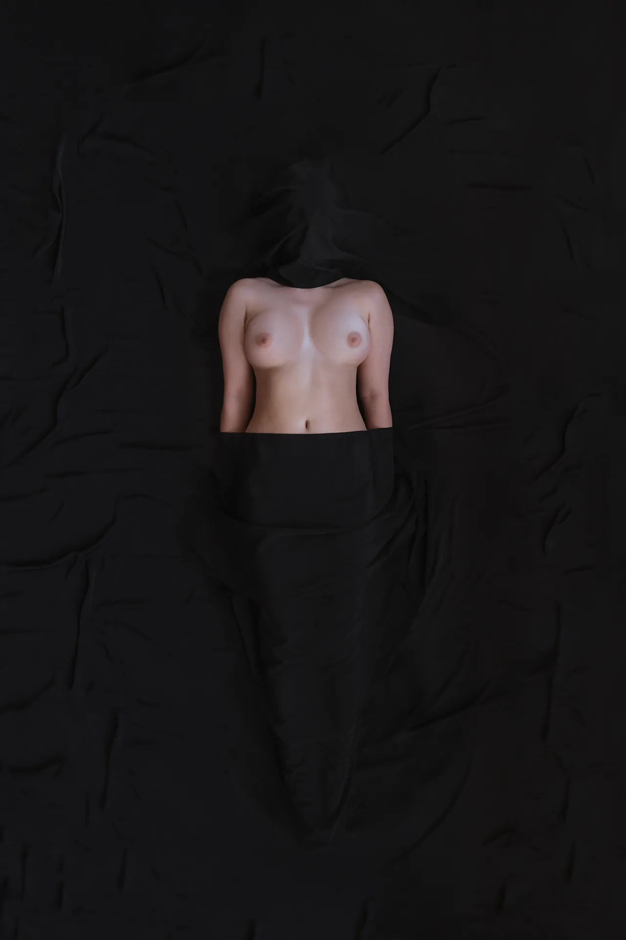 A bare torso emerges from folds of black fabric against a nearly black background.