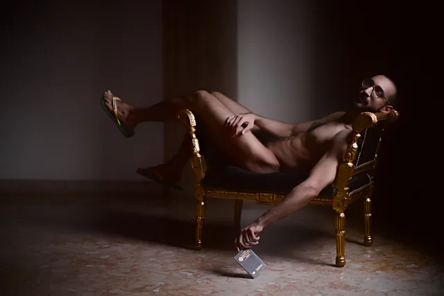 A nude figure reclines on an ornate gold chair, wearing bright sandals, with one hand lowering a small dark booklet or document toward the floor; no text is readable on it.