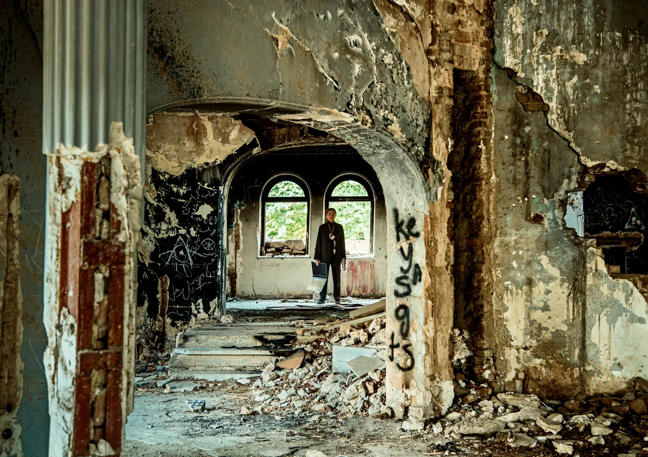 A person stands framed by successive arched doorways inside a heavily damaged building.