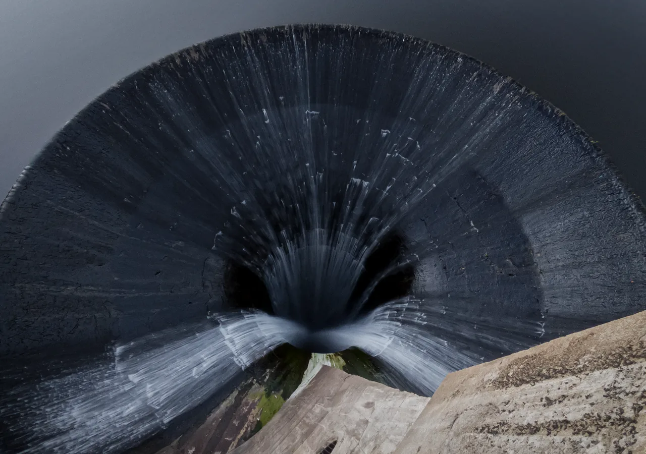 The camera looks upward through a dark, fluted circular structure toward a small opening of sky.