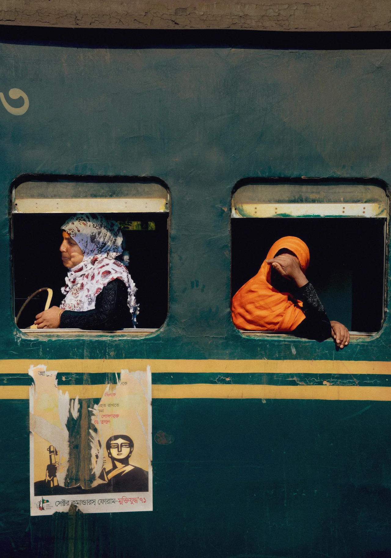 Two people lean from the windows of a weathered teal train; one wears a bright orange head covering.