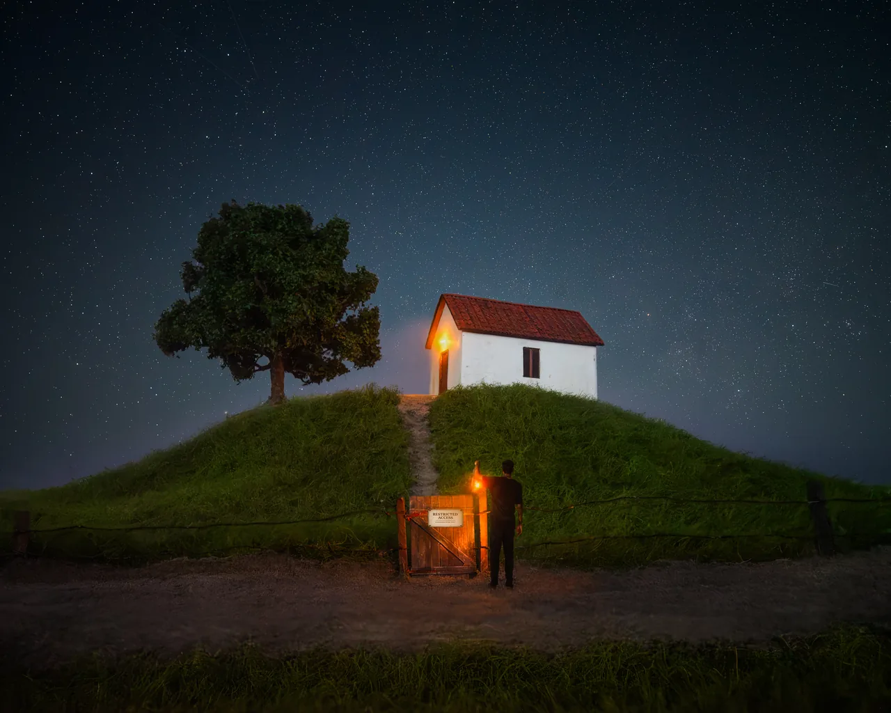 A small white house with a red roof stands on a hill beneath a starry sky, beyond a lit gate with a posted sign and a silhouetted figure.