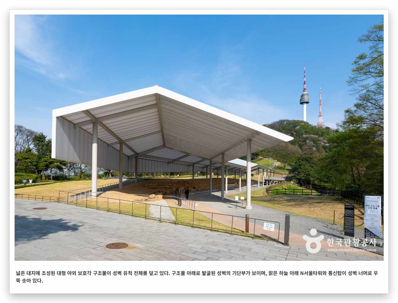 넓은 대지에 조성된 대형 야외 보호각 구조물이 성벽 유적 전체를 덮고 있다. 구조물 아래로 발굴된 성벽의 기단부가 보이며, 맑은 하늘 아래 N서울타워와 통신탑이 성벽 너머로 우뚝 솟아 있다.