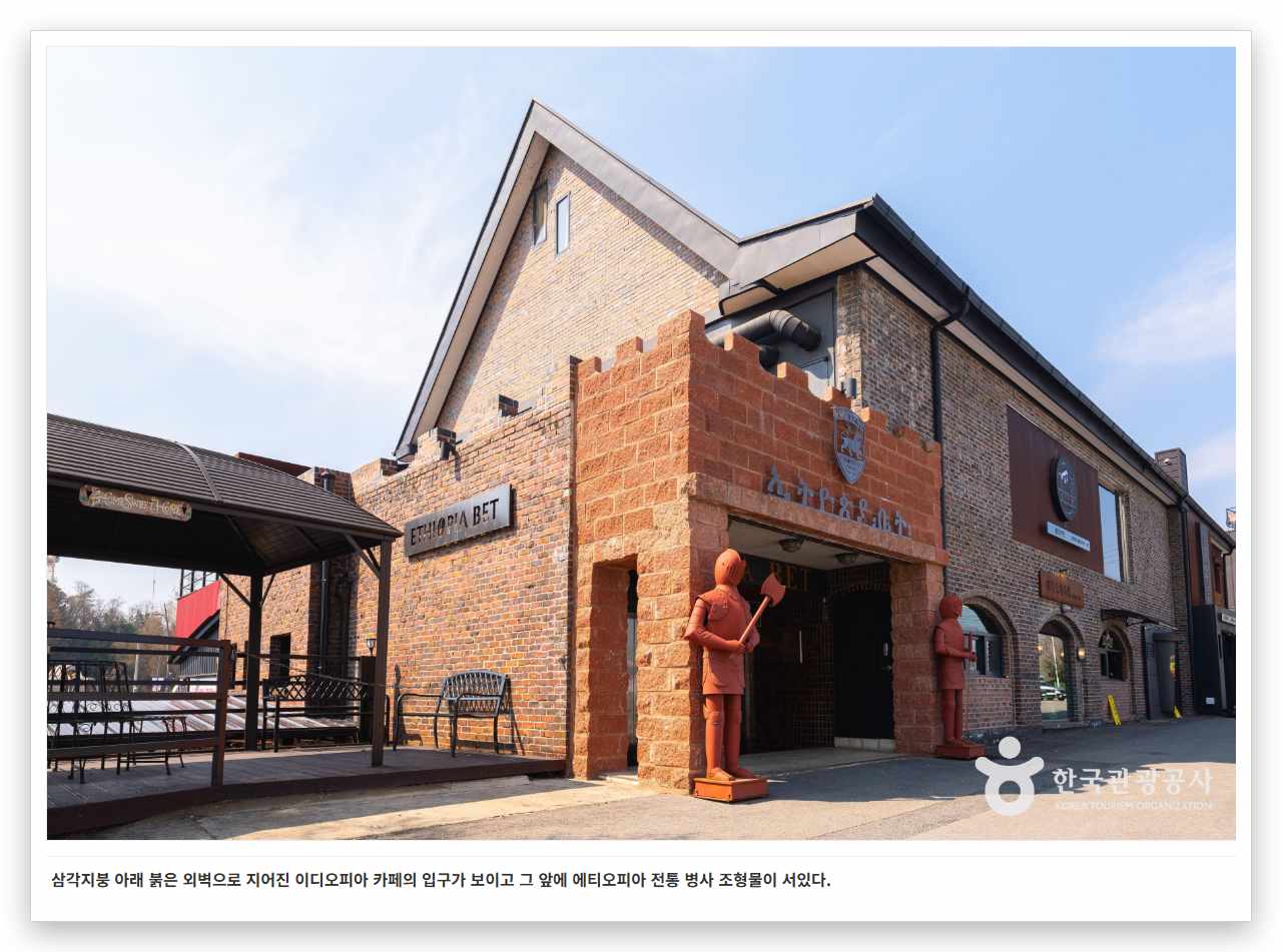 삼각지붕 아래 붉은 외벽으로 지어진 이디오피아 카페의 입구가 보이고 그 앞에 에티오피아 전통 병사 조형물이 서있다. 