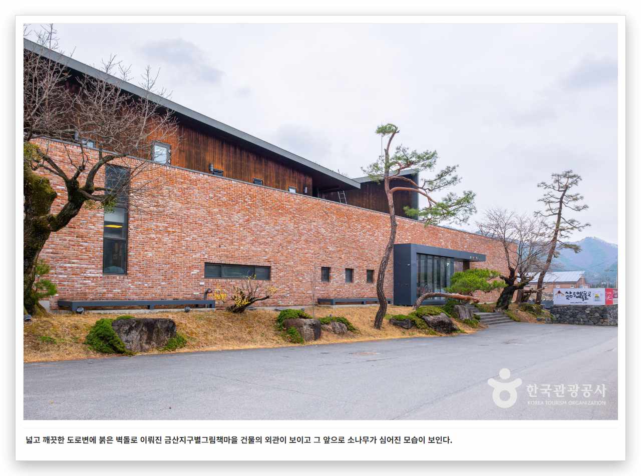 넓고 깨끗한 도로변에 붉은 벽돌로 이뤄진 금산지구별그림책마을 건물의 외관이 보이고 그 앞으로 소나무가 심어진 모습이 보인다.