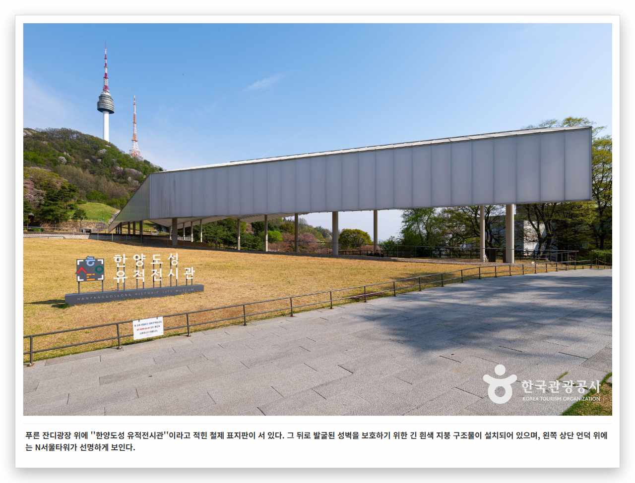 푸른 잔디광장 위에 ''한양도성 유적전시관''이라고 적힌 철제 표지판이 서 있다. 그 뒤로 발굴된 성벽을 보호하기 위한 긴 흰색 지붕 구조물이 설치되어 있으며, 왼쪽 상단 언덕 위에는 N서울타워가 선명하게 보인다.