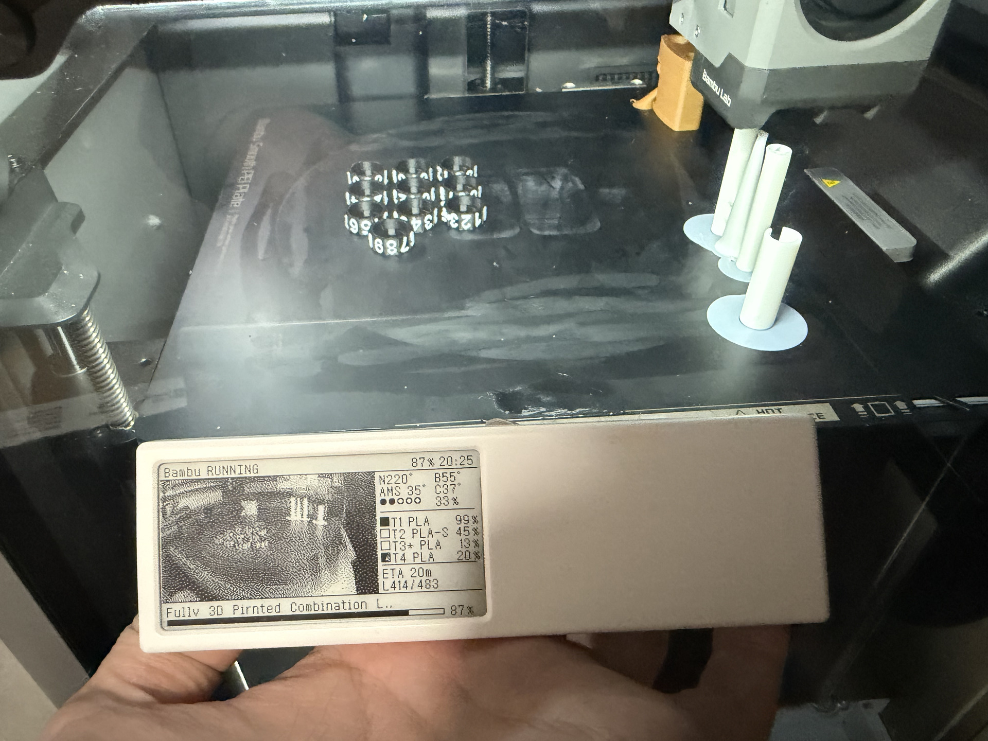 A hand-held Quote/0 showing printer status in front of the Bambu Lab printer and its printed parts