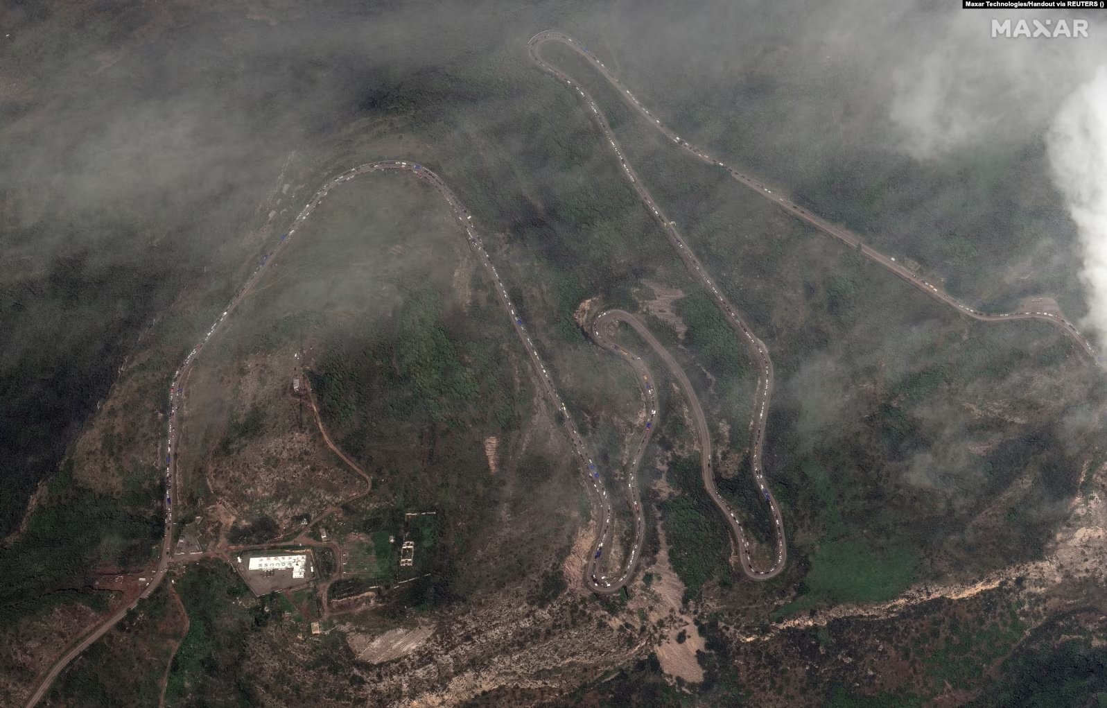 An aerial view of the Stepanakert-Goris roadway connecting Artsakh to Armenia via the Lachin Corridor. A lone roadway winds between mountainous landscapes; the entirety of the road is dotted with rows of vehicles.