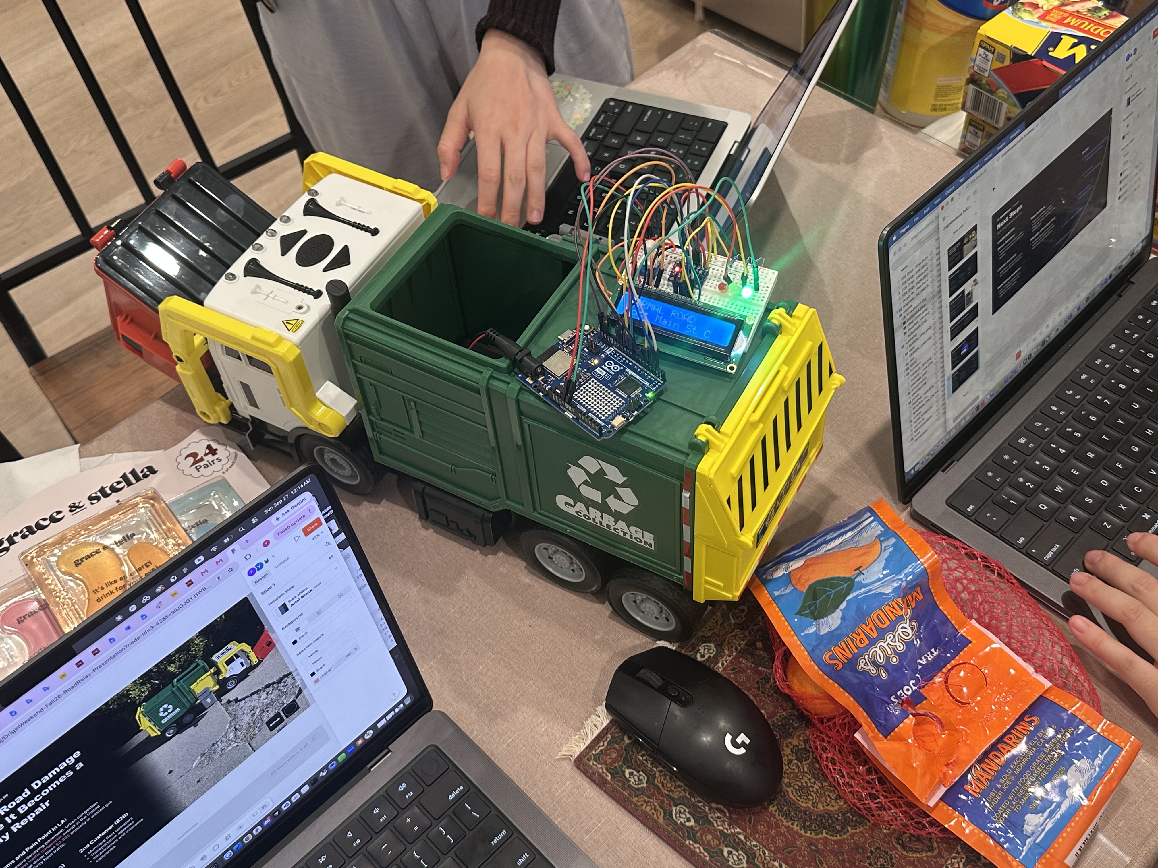 RoadRelay work in progress: the Arduino, sensor breadboard, LCD, and LEDs mounted on a toy garbage truck beside the team's laptops
