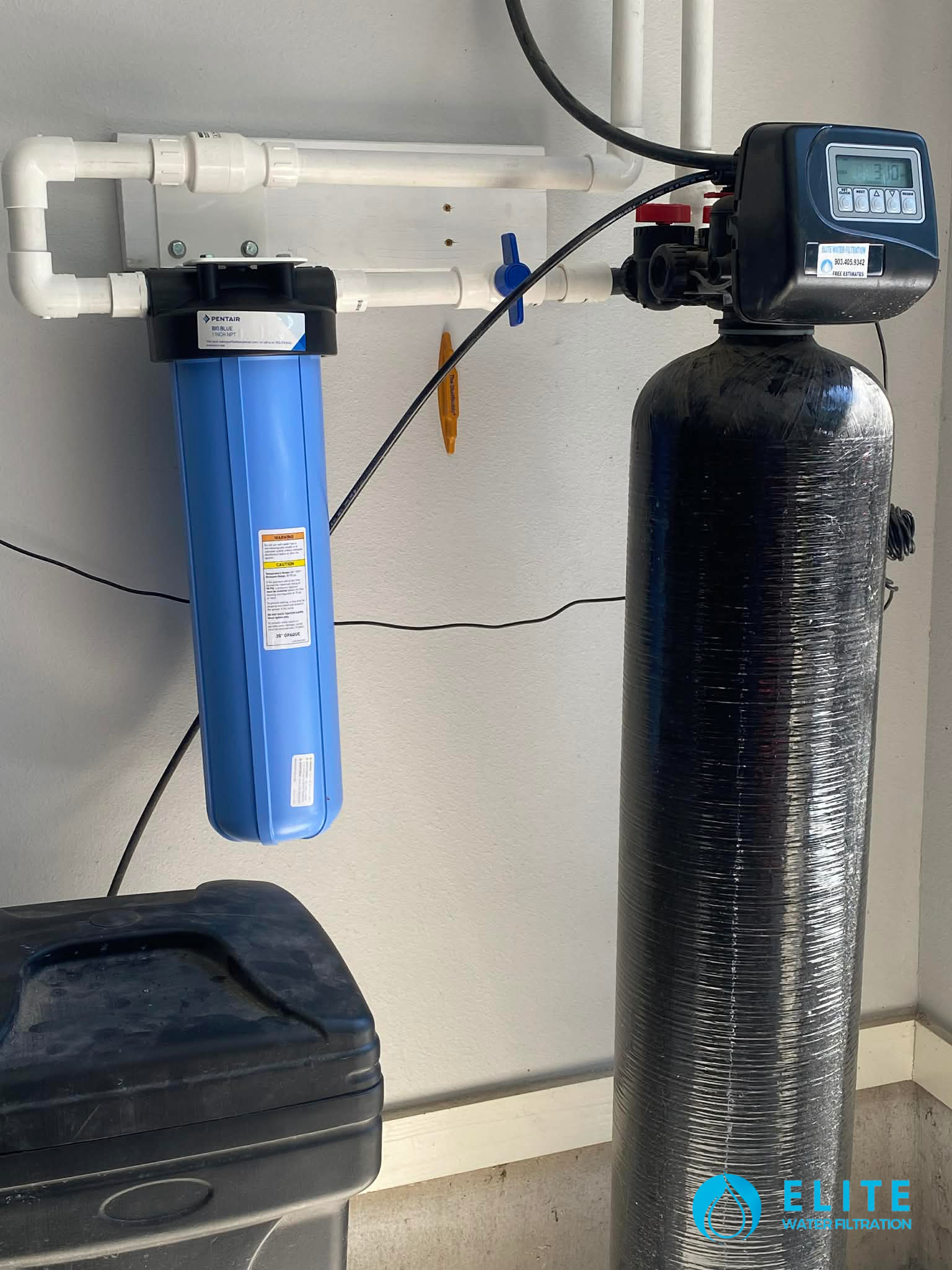 Installed water softener tank with electronic head, mounted on a white wall with PVC plumbing.