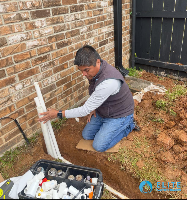 Worker installing PVC piping in an outdoor trench next to a brick house.