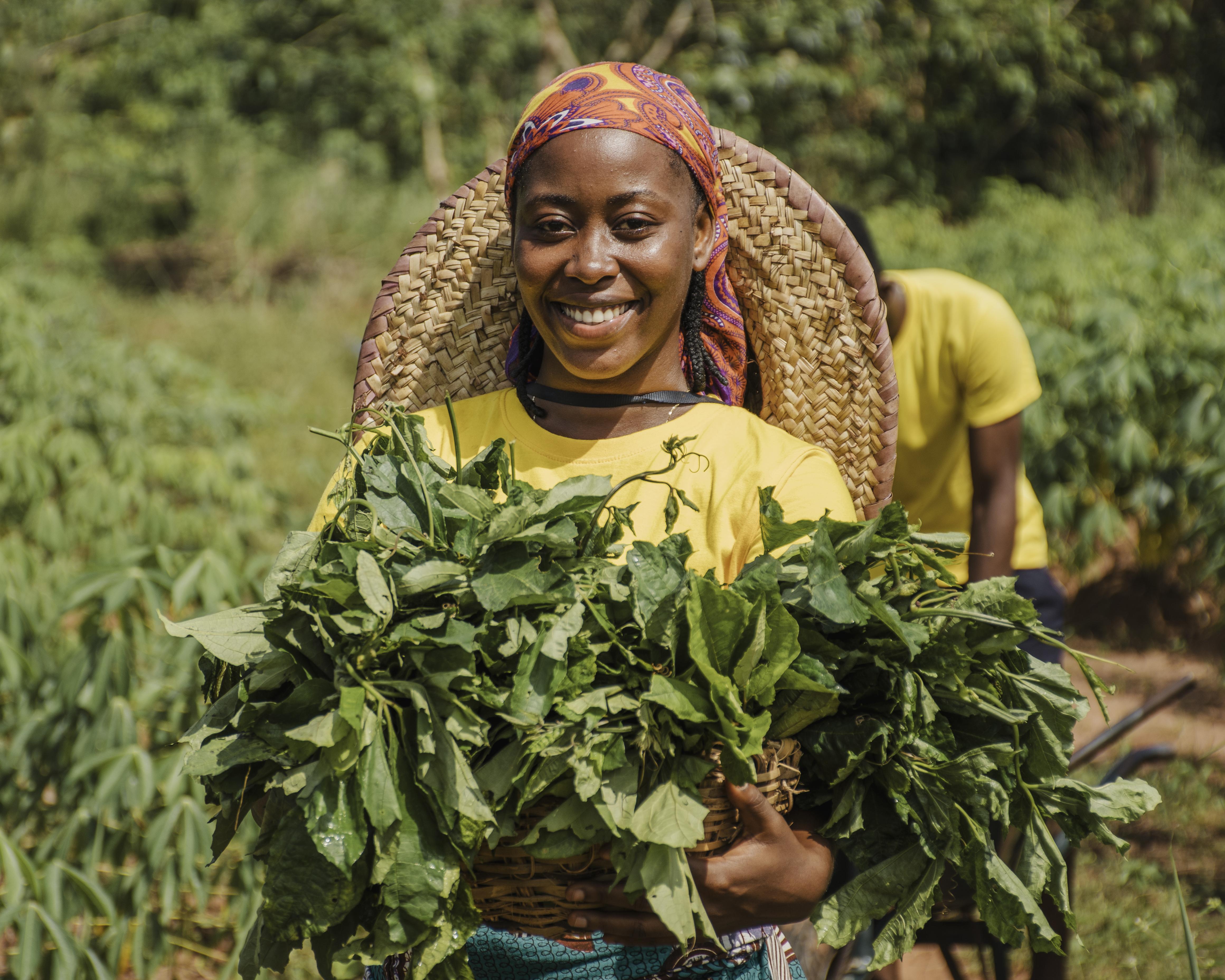 A smiling black woman with an orange does and yellow shirt holding a basket with crops. In the background is a farm with someone working.