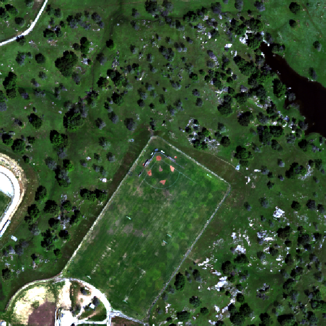 RGB image of the SJER field site. At the top right of the image, there is dark, brakish water. Scattered throughout the image, there are several trees. At the center of the image, there is a baseball field, with low grass. At the bottom left of the image, there is a parking lot and some buildings with highly reflective surfaces, and adjacent to it is a section of a gravel lot.