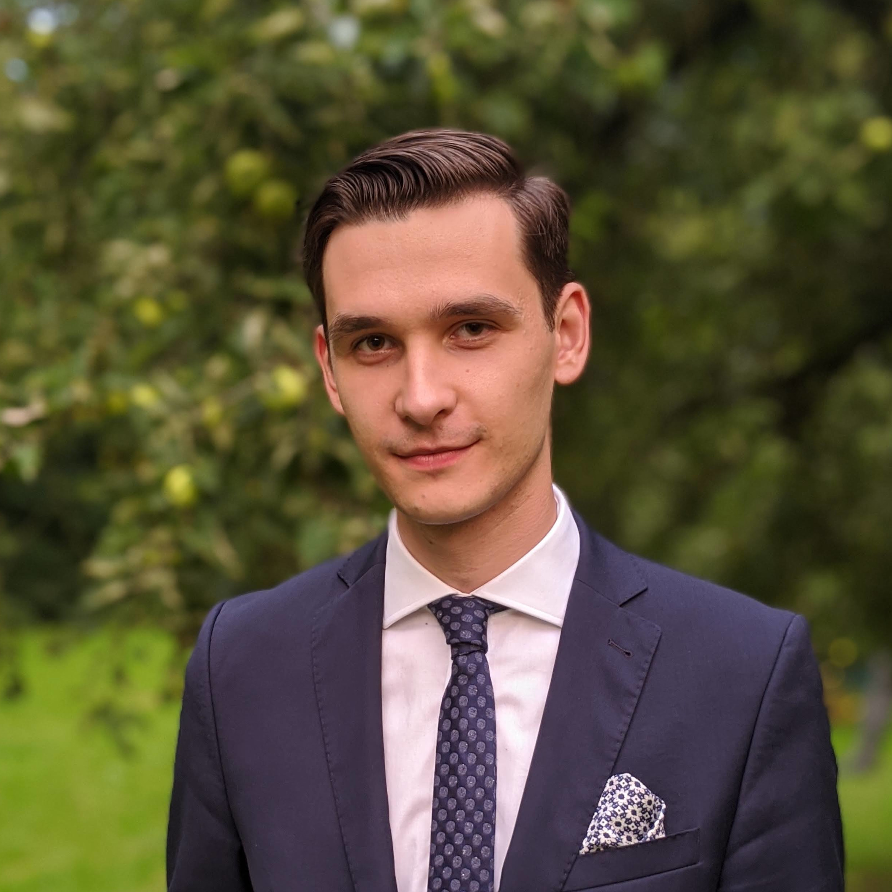 image of Michal Sadowski wearing a navy suit in a dim nature background