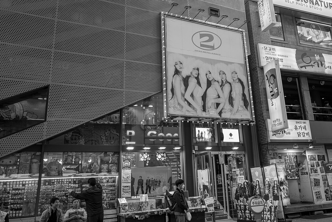 A Seoul street in May 2024, with a large (G)I-DLE second full album billboard mounted above a shop and Korean signage on every storefront. Photo by David Lee from Redmond, WA, USA, CC BY 2.0, via Wikimedia Commons