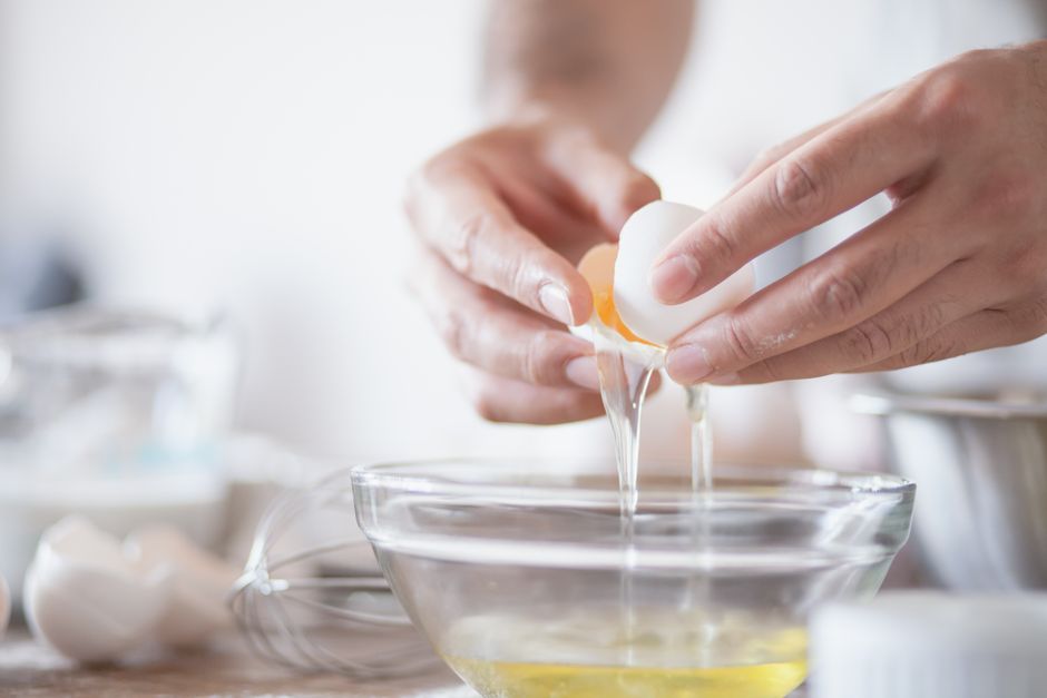 A close-up of hands cracking a raw egg over a glass mixing bowl in a home kitchen, egg white already dripping in