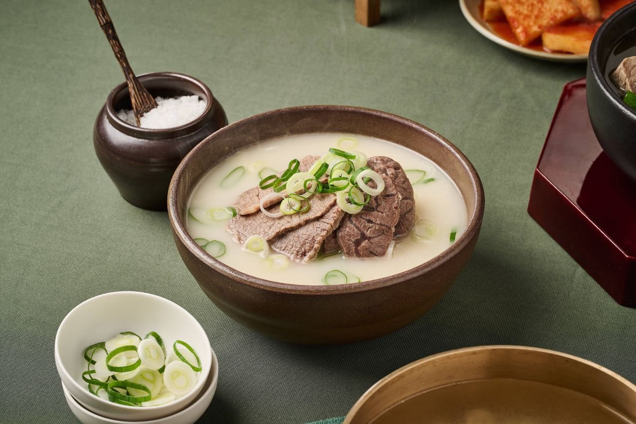 A bowl of seolleongtang milky beef bone broth topped with sliced beef and scallions, served with a small dish of salt and scallions on the side