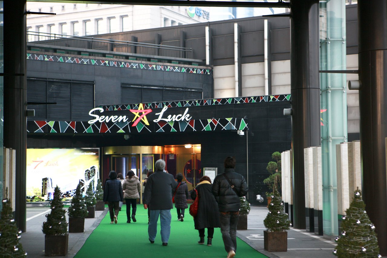 Seoul's Seven Luck casino at dusk, its lit sign above a green carpet leading toward the entrance