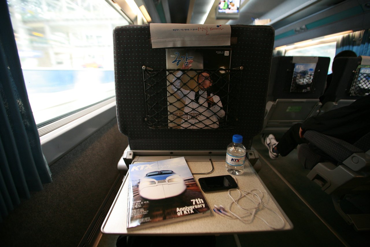 A KTX tray table with the onboard magazine, a KTX-branded water bottle and a phone, the seat-back pocket behind it. ⓒ한국관광공사 포토코리아-이범수 (Korea Tourism Organization, KOGL Type 1)