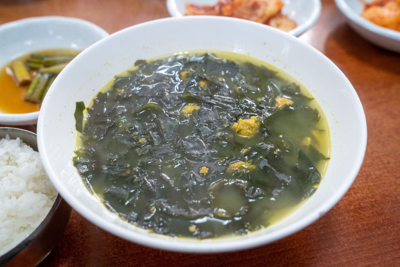 A white bowl of miyeokguk, Korean seaweed soup, with rice and side dishes beside it