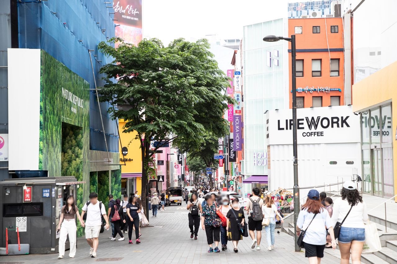 A busy Myeongdong shopping street in Seoul lined with cosmetics and coffee shop signage, crowded with pedestrians on a weekday afternoon — ⓒ한국관광공사 포토코리아-이범수 (Korea Tourism Organization, KOGL Type 1)