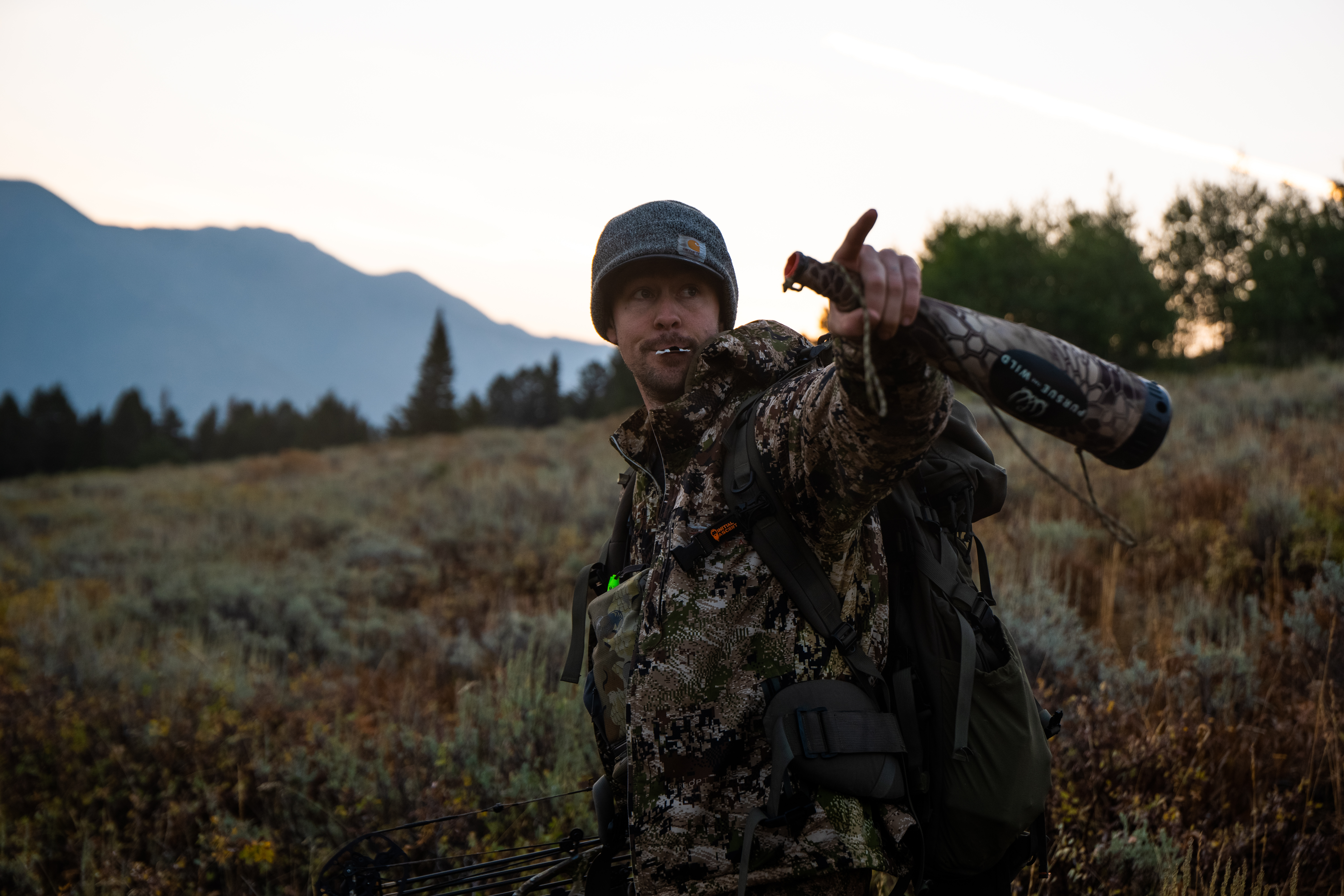 Zach in camo pointing across a mountain meadow at dusk