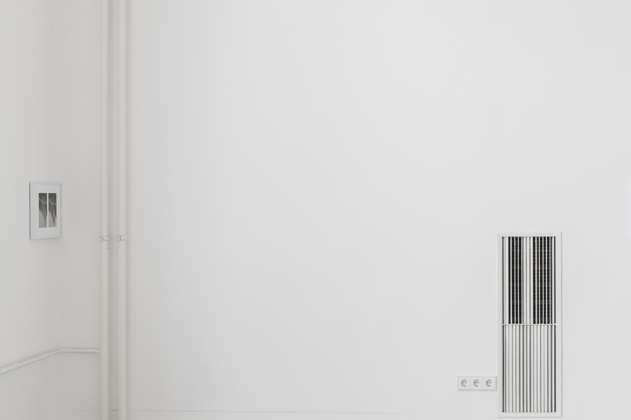 Aisha Altenhofen, The Carrier at UdK Berlin: Side profile of a dual-photo frame positioned near white vertical pipes and an expansive empty wall section.