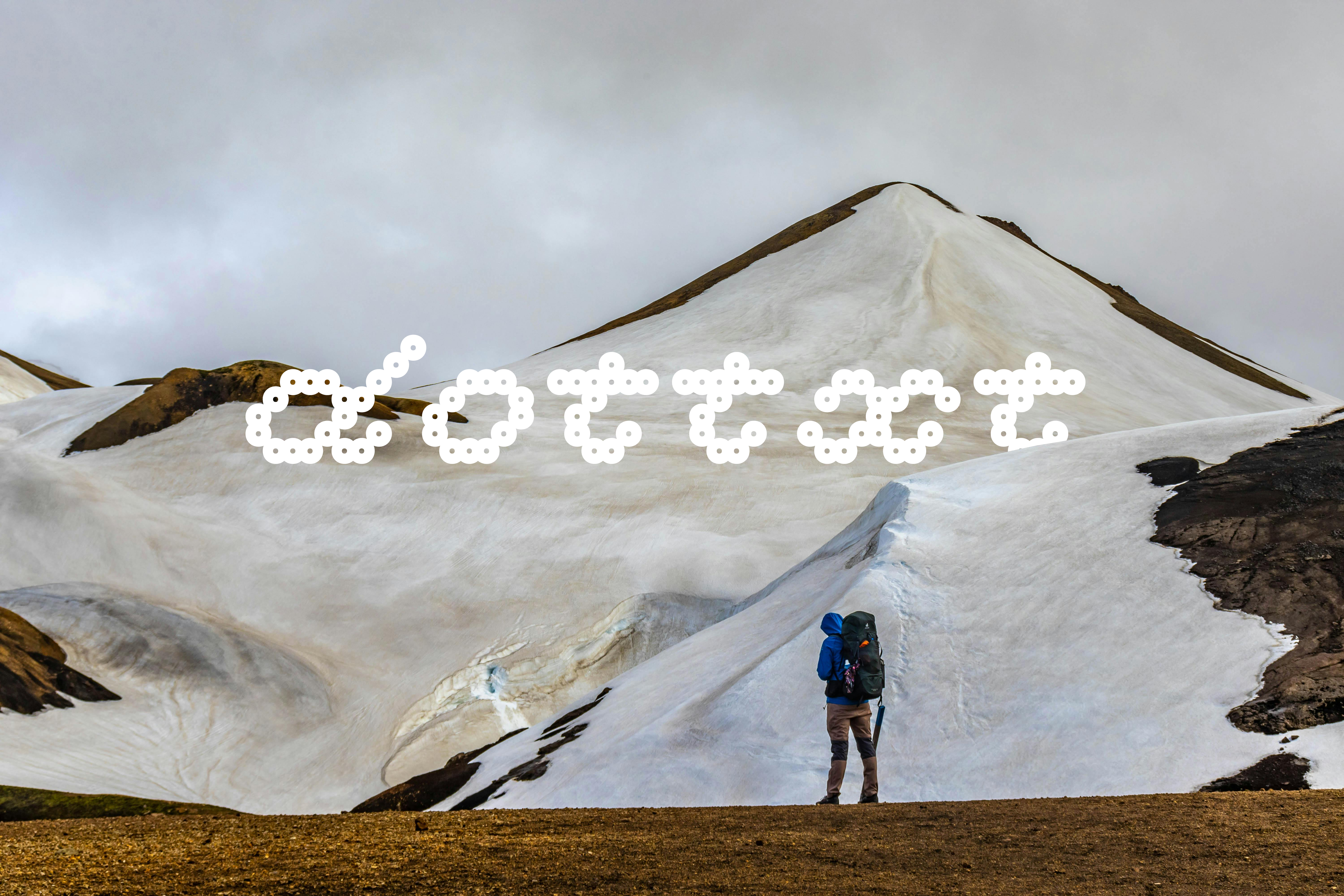 a person with a large backpack, heavy blue jacket, beige snow pants, and snow boots standing right in front of mountains covered in snow. between the mountains is the text dottxt in a decorative font, with the text being partiallly obscured by the mountains.