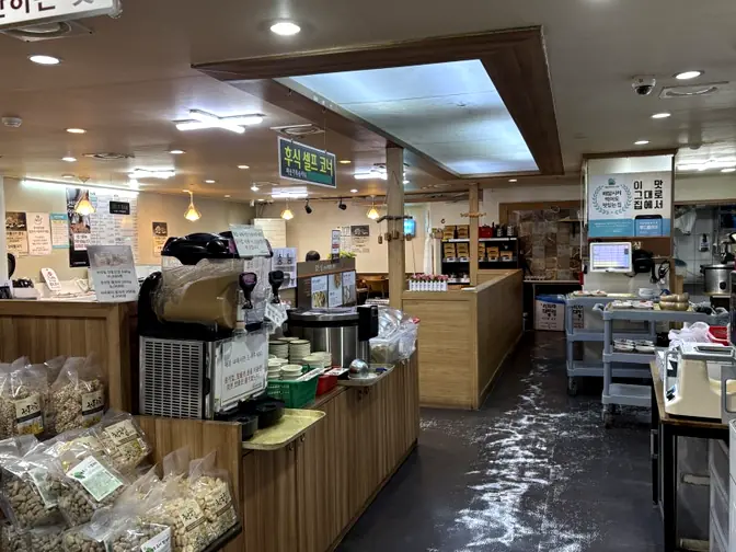 A wide interior view of the Cheonggukjang and Boribap Cheongdam restaurant, showcasing the checkout counter, various packaged food items for sale, and the overall rustic-modern ambiance with warm lighting.