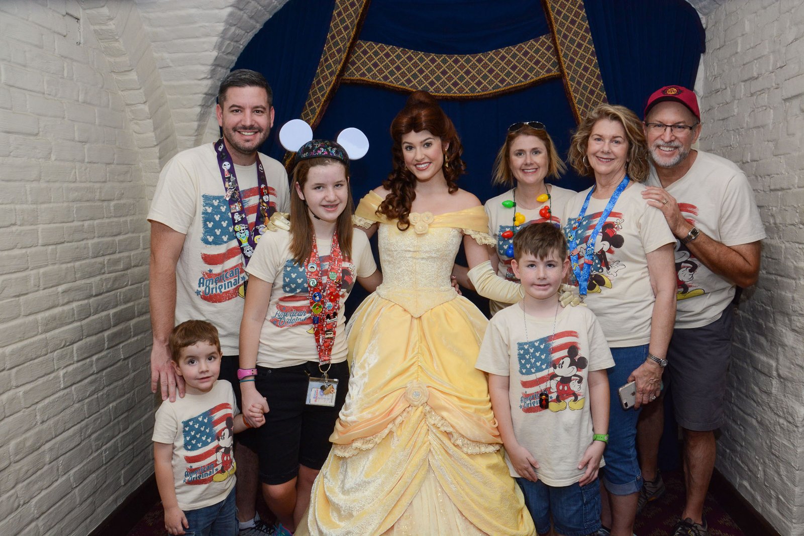 A family poses with Belle inside Akershus Royal Banquet Hall at EPCOT's Norway pavilion, Walt Disney World — Belle in her yellow ballgown in the restaurant's greeting alcove, with blue drapes and white brick walls behind them.