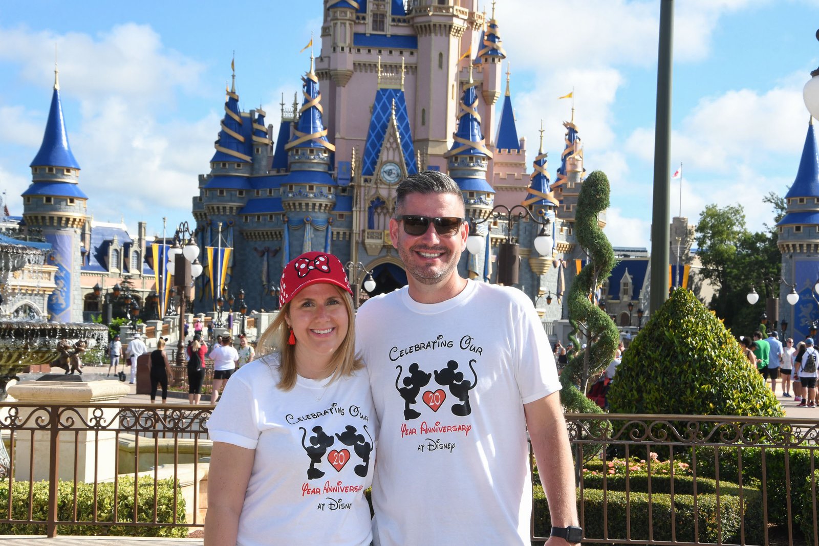Cinderella Castle at Magic Kingdom in its 50th-anniversary paint — pink walls, royal-blue turrets and gold ribbons — with a couple posing in front of it on a sunny day at Walt Disney World.