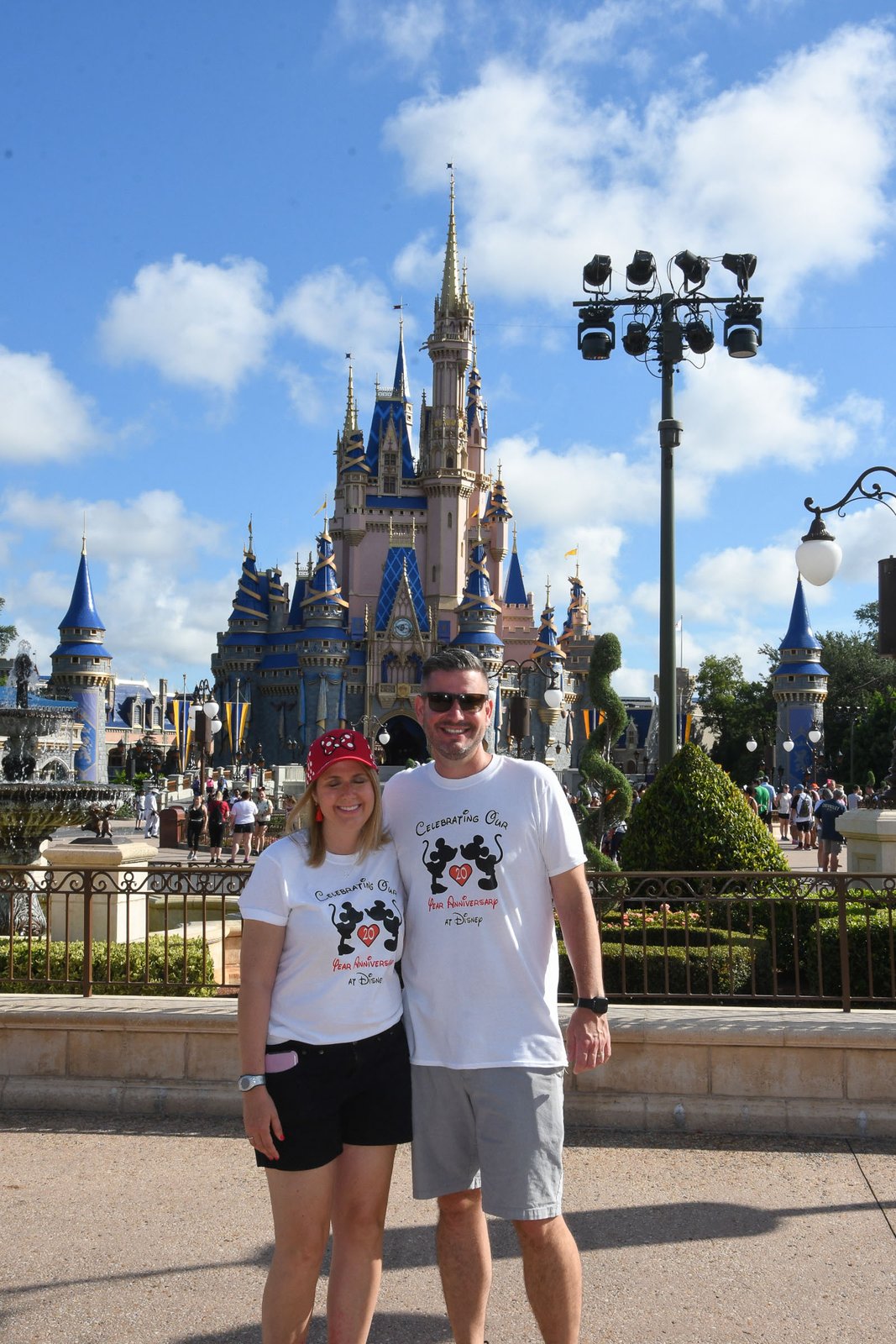 Cinderella Castle in bright July sunshine at Magic Kingdom — blue sky, white clouds and summer crowds around the hub, with a couple posing in front of the castle.