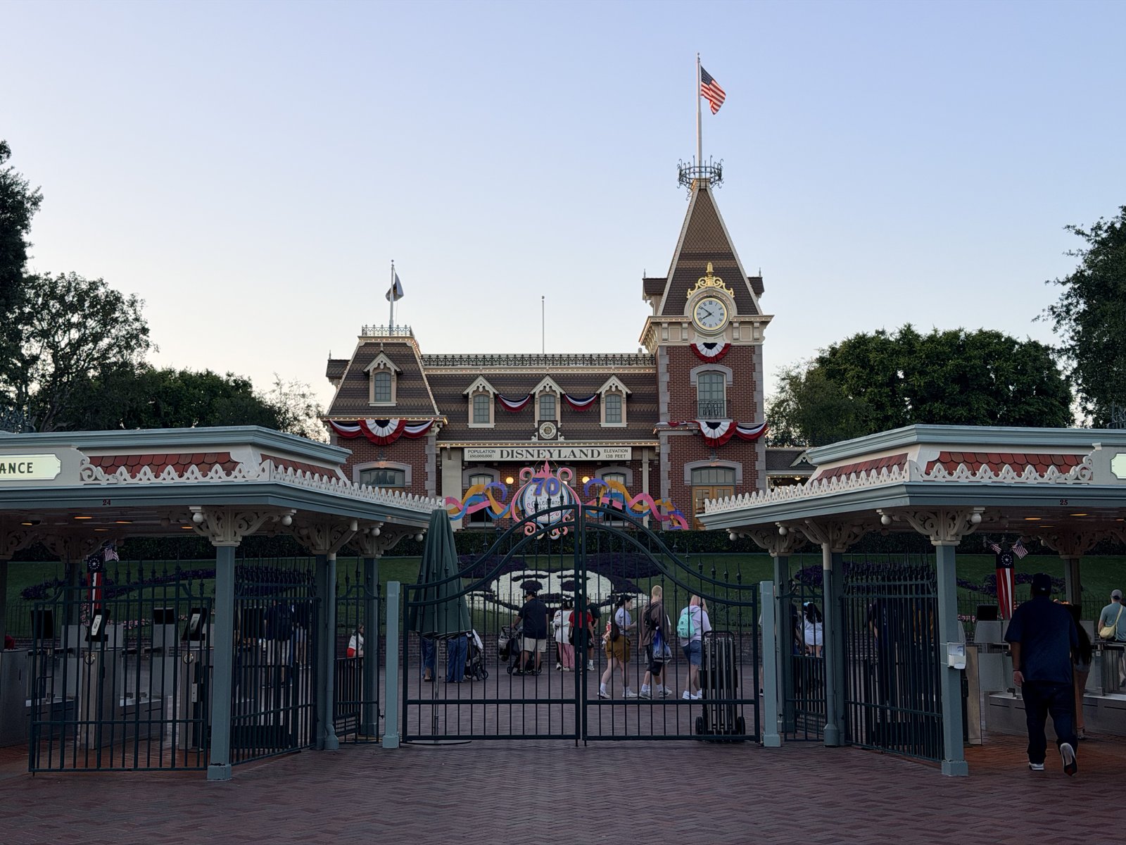 Disneyland's entrance gates and Main Street train station at dusk, with the DISNEYLAND sign and the floral Mickey Mouse display on the hillside below the station.