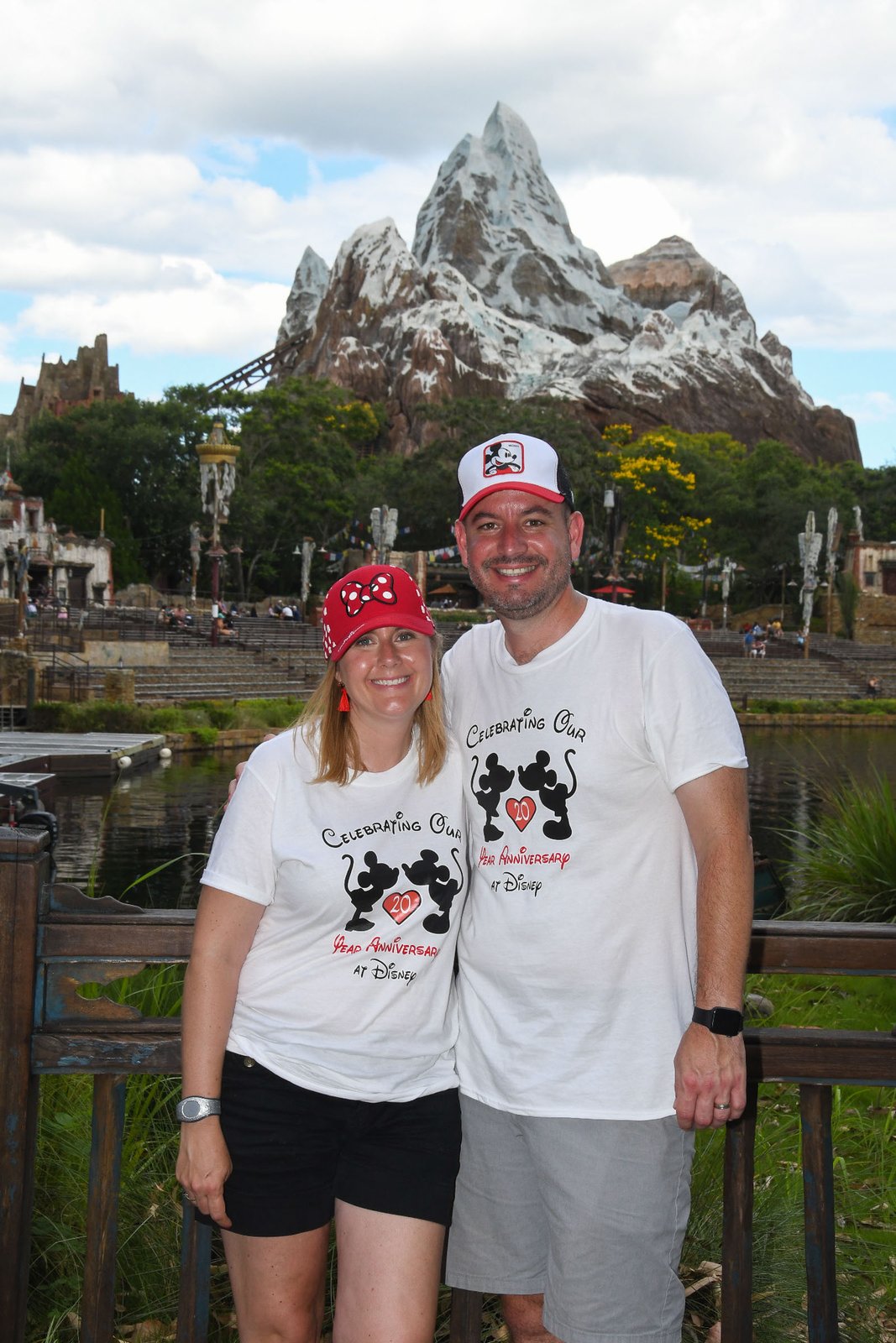 Expedition Everest rises across Discovery River at Disney's Animal Kingdom, its snow-capped Himalayan peak above the riverside seating terraces, with a couple posing on the bridge in the foreground.