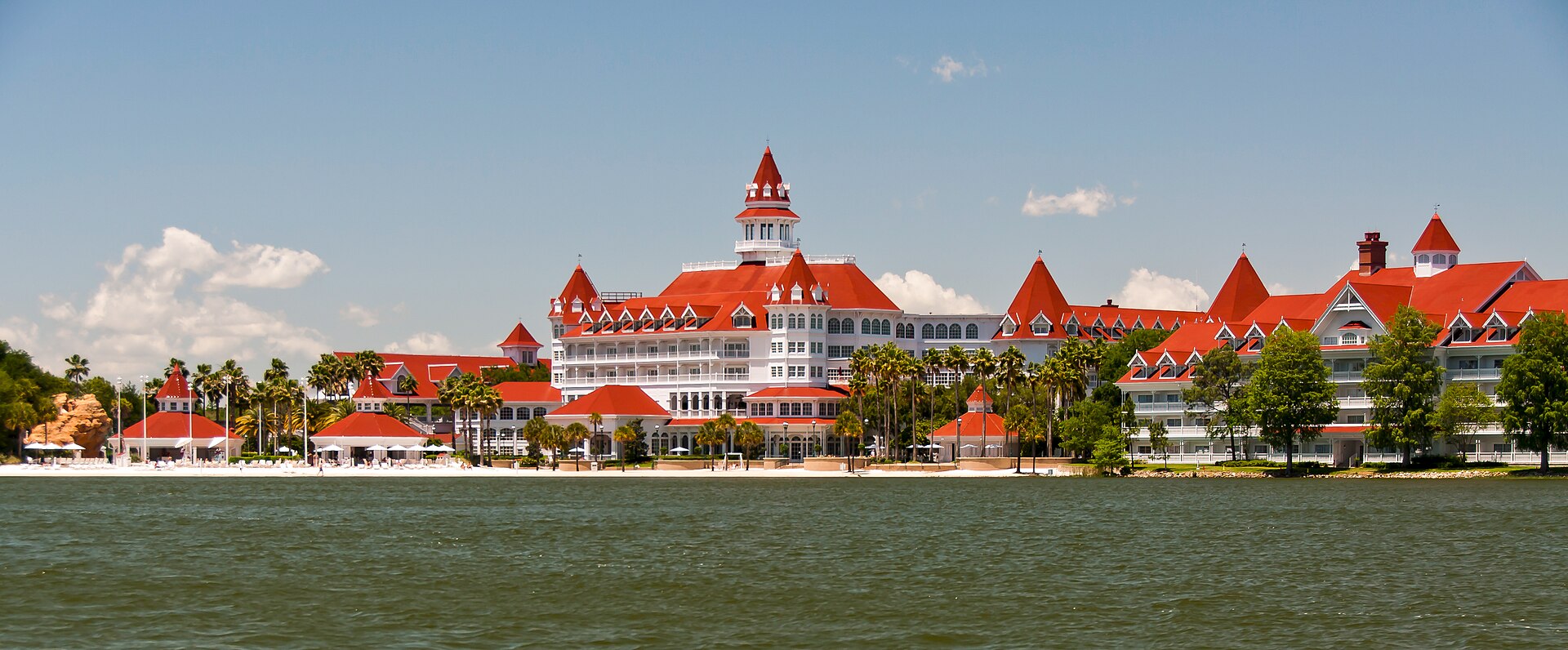 Disney's Grand Floridian Resort & Spa seen across Seven Seas Lagoon at Walt Disney World — red roofs and a white Victorian facade above the beach and palm trees.
