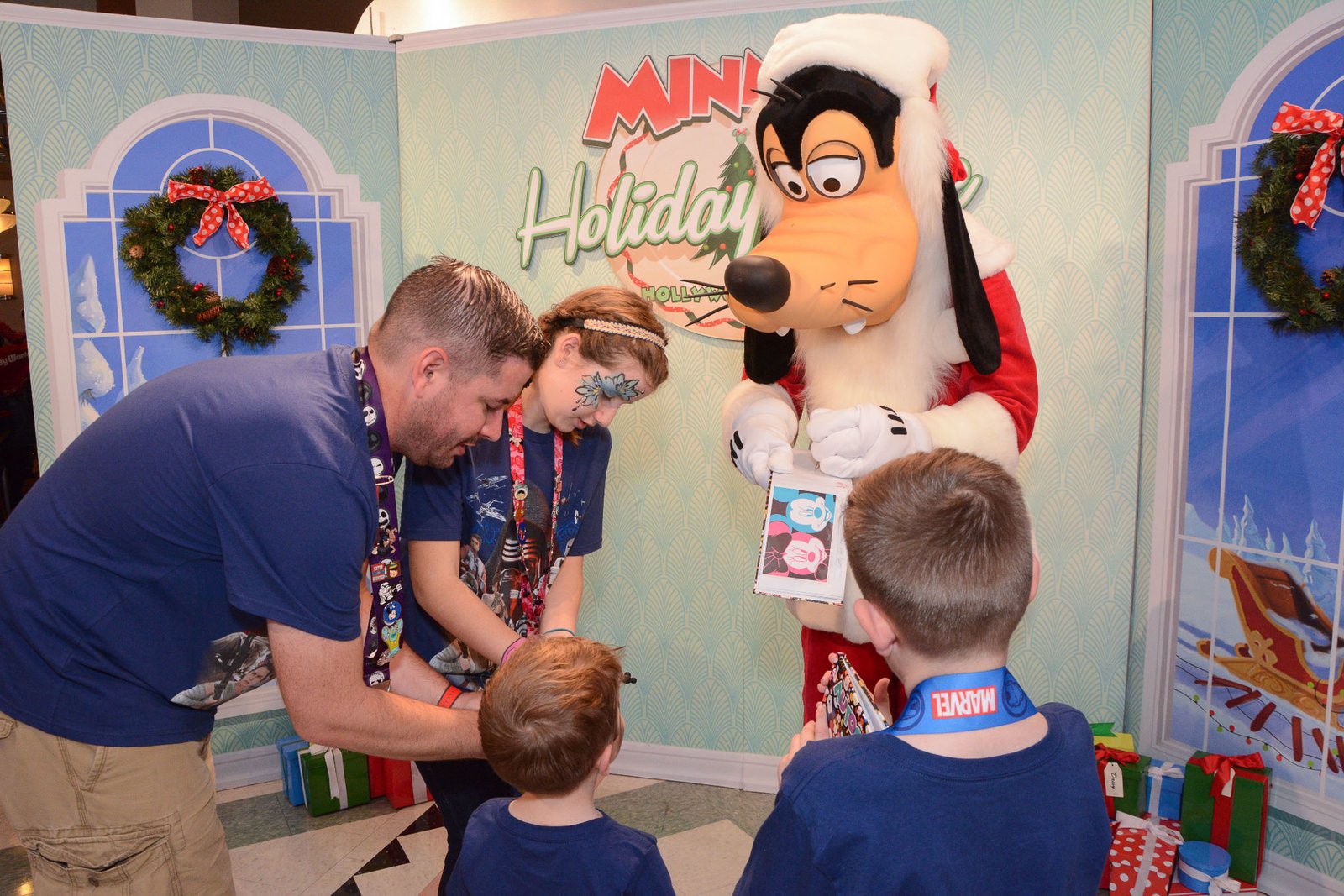 Santa Goofy signs an autograph book for children at Minnie's Holiday Dine at Hollywood & Vine in Disney's Hollywood Studios, in front of the Christmas backdrop with wreaths and wrapped gifts.