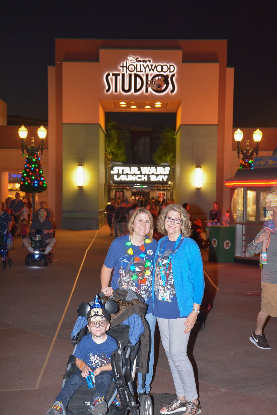 The Disney's Hollywood Studios entrance arch lit up at night, its sign glowing above the walkway with the Star Wars Launch Bay sign beyond, and a family posing in front.
