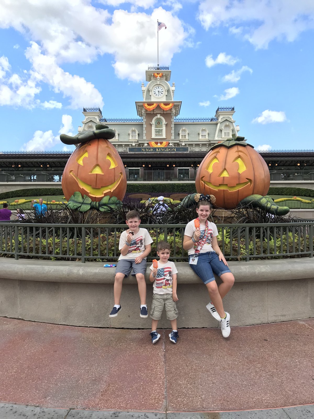 Three kids eat ice pops on a planter wall in front of the giant jack-o'-lantern topiaries at the Magic Kingdom entrance, with the Main Street train station behind them dressed for the fall Halloween season at Walt Disney World.