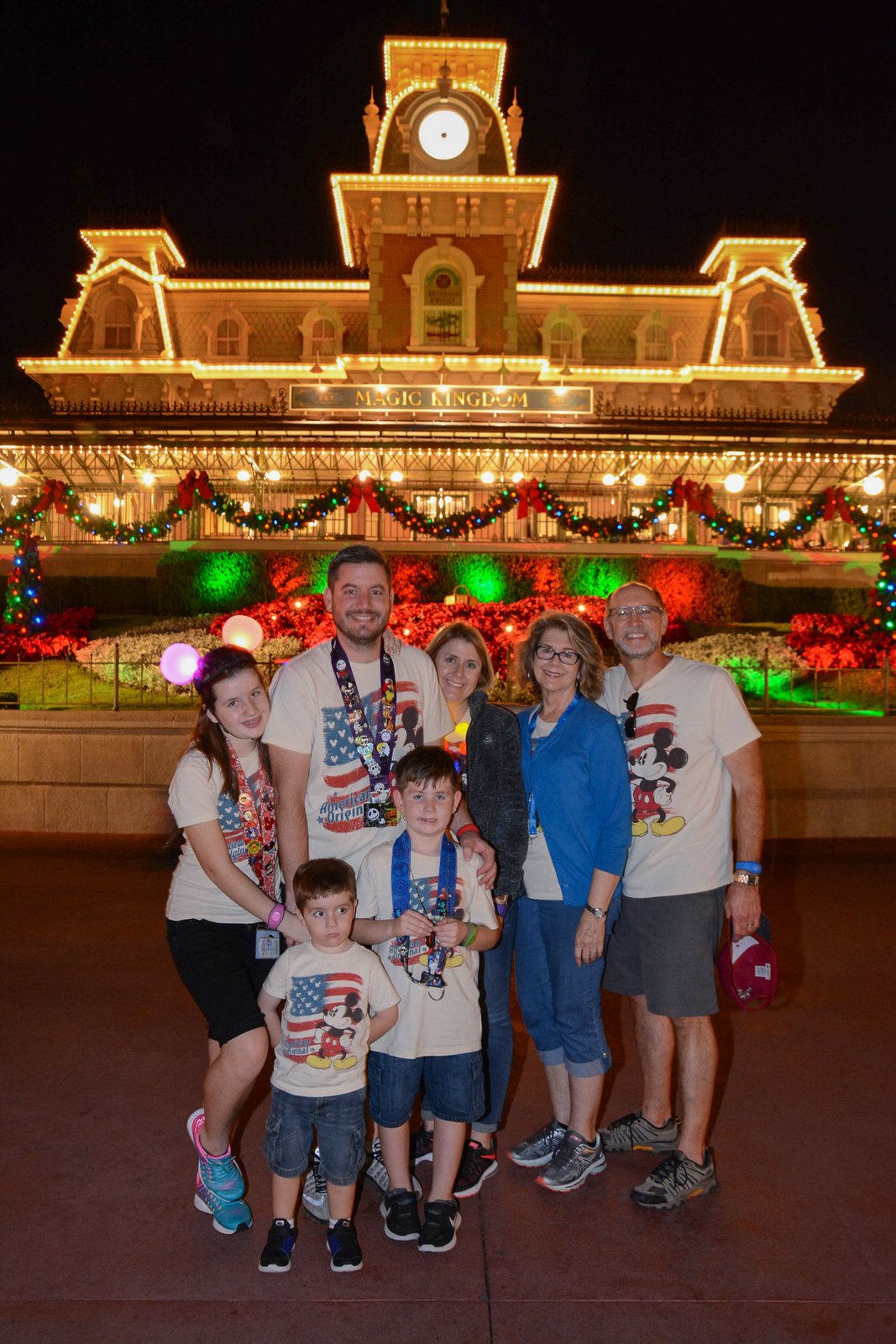 The Magic Kingdom train station glows at night in the Christmas season, outlined in warm lights with garlands, red bows and poinsettias across the park entrance, and a family posing in front at Walt Disney World.