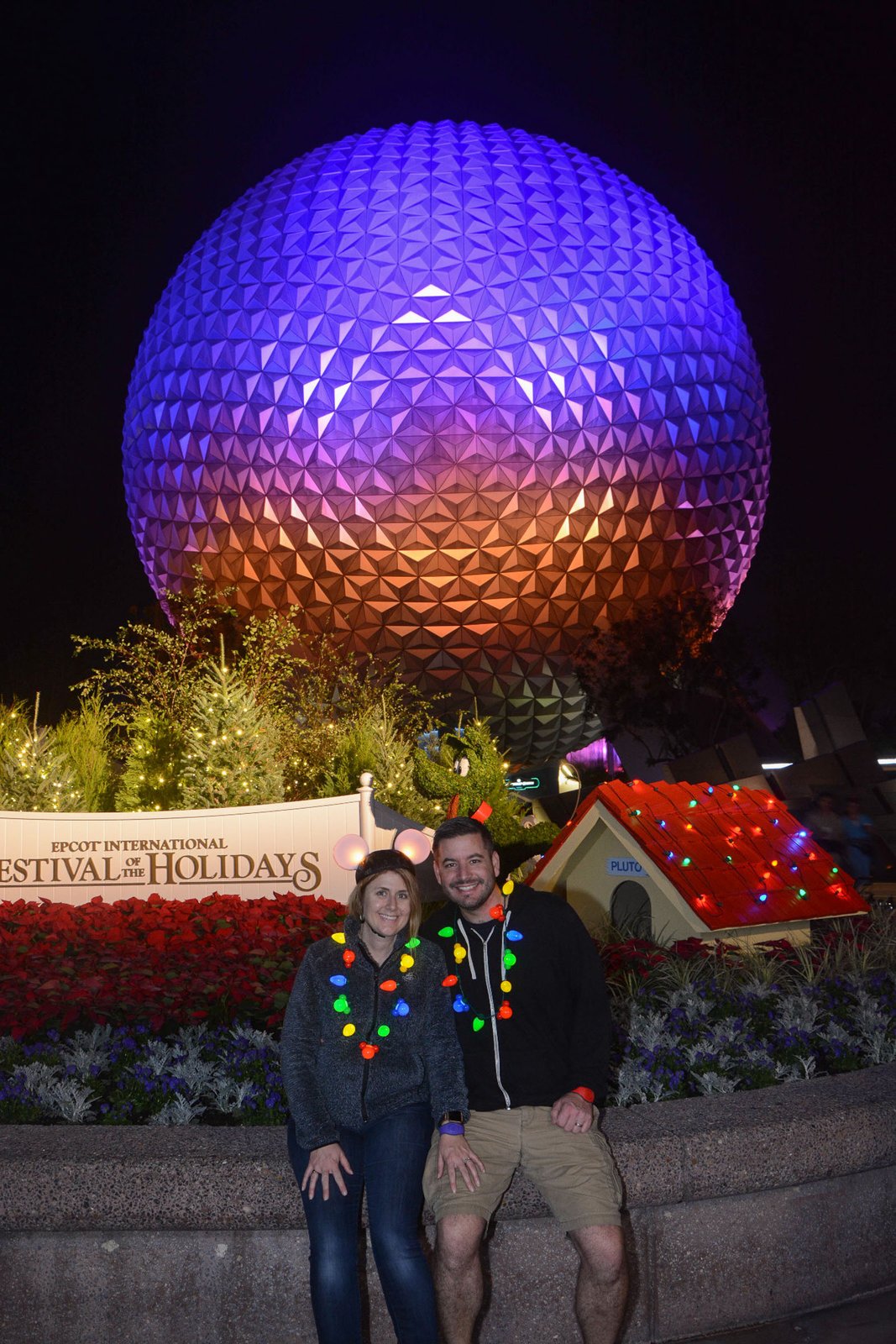 Spaceship Earth glows purple and orange above the EPCOT International Festival of the Holidays sign at night, with poinsettias, small lit Christmas trees and Pluto's doghouse in the foreground at EPCOT.