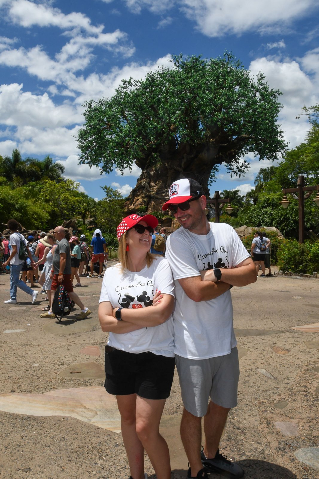 A couple laughs together in front of the Tree of Life on Discovery Island at Disney's Animal Kingdom, its carved trunk and green canopy rising behind them on a sunny day.