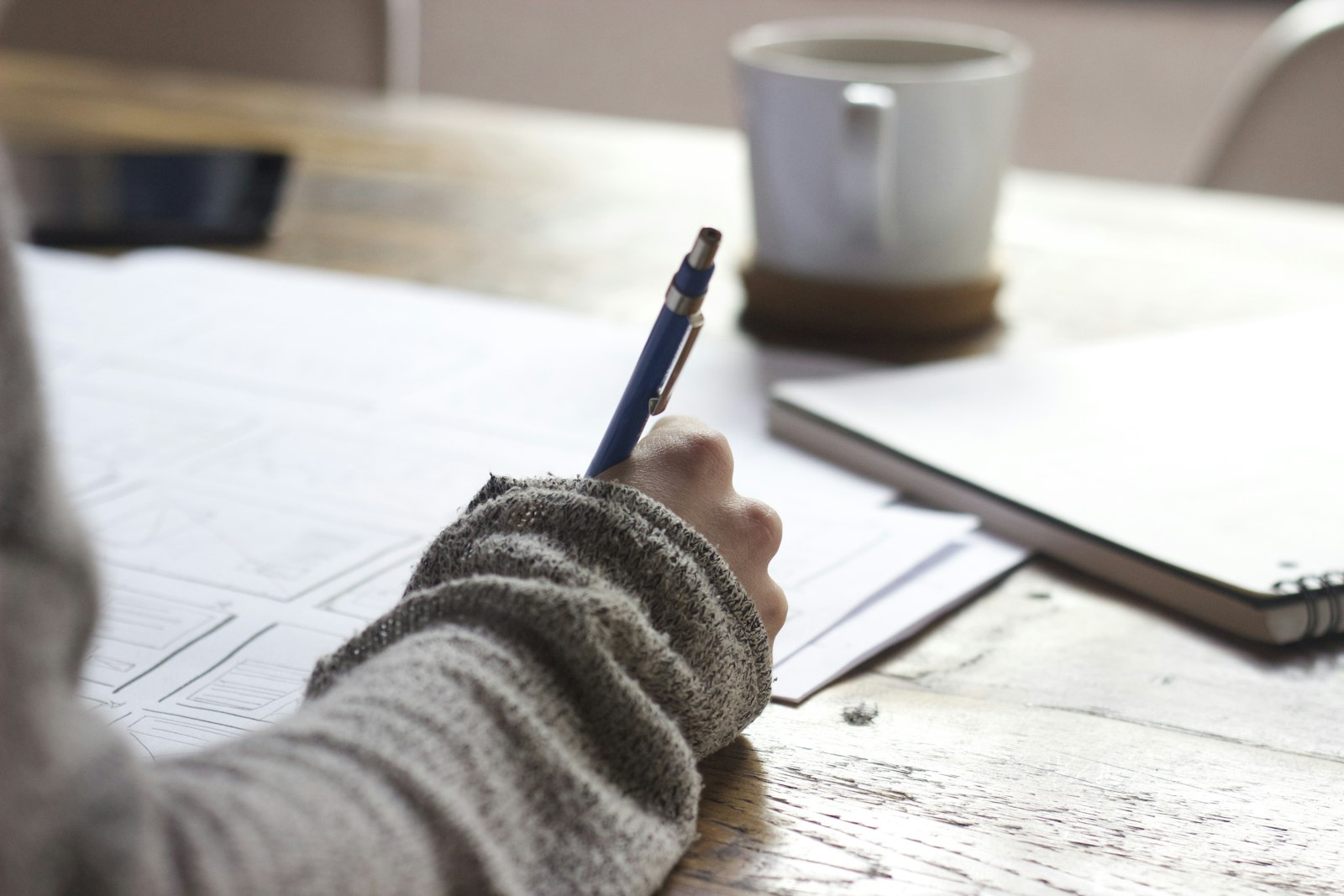 Person writing notes in a notebook on a wooden desk beside a coffee mug