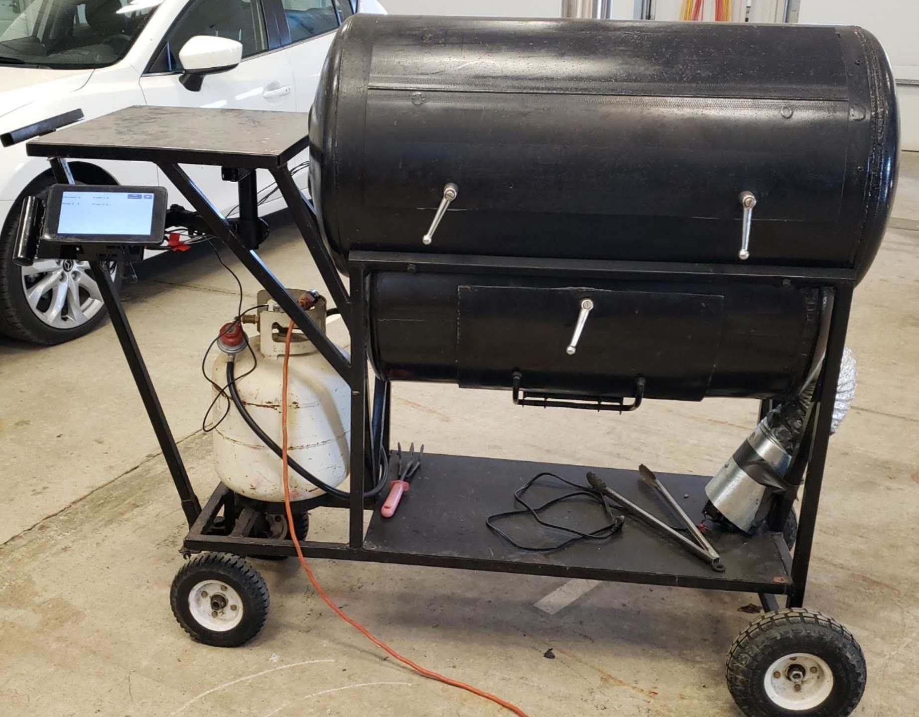 The pit itself — a barrel smoker on a welded steel cart, with the Raspberry Pi touchscreen kiosk mounted on the side shelf running the smoker app, the propane burner below it and the blower assembly at the far end