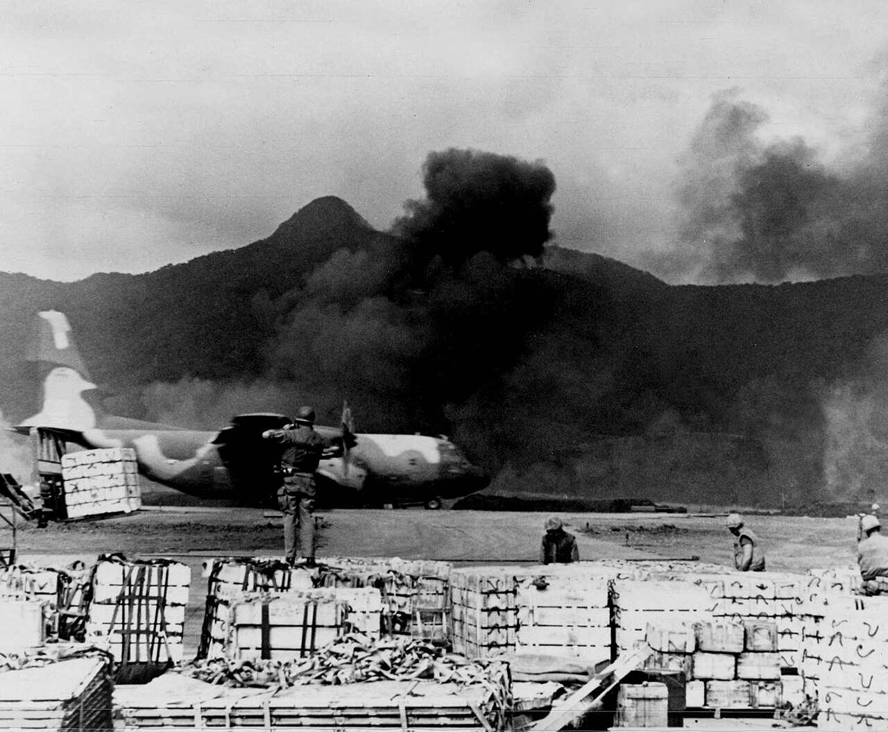A C-130 on the Khe Sanh airstrip amid supply pallets, smoke, and the surrounding hills, 1968