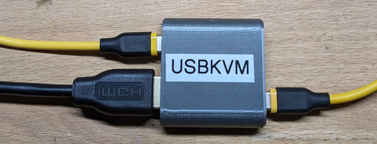 A grey, obround 3D-printed 5x5 cm box with a white "USBKVM" label on it. A black HDMI cable an a yellow USB Type-C cable are connected on the left side. Another yellow USB Type-C cable is plugged into the right side.